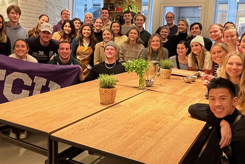 Accounting Study Abroad group gathers around a large table, some holding up a large TCU flag