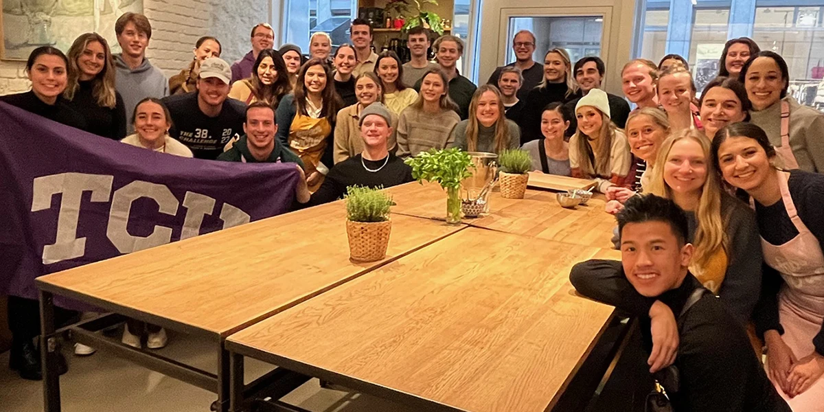 Accounting Study Abroad group gathers around a large table, some holding up a large TCU flag